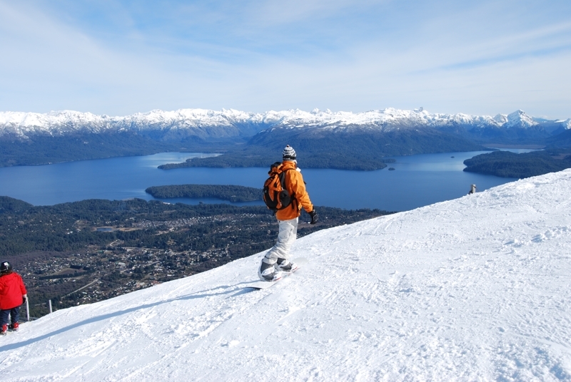 persona practicando Snowboard con paisaje de fondo compuesto por lago, pueblo y montañas nevadas