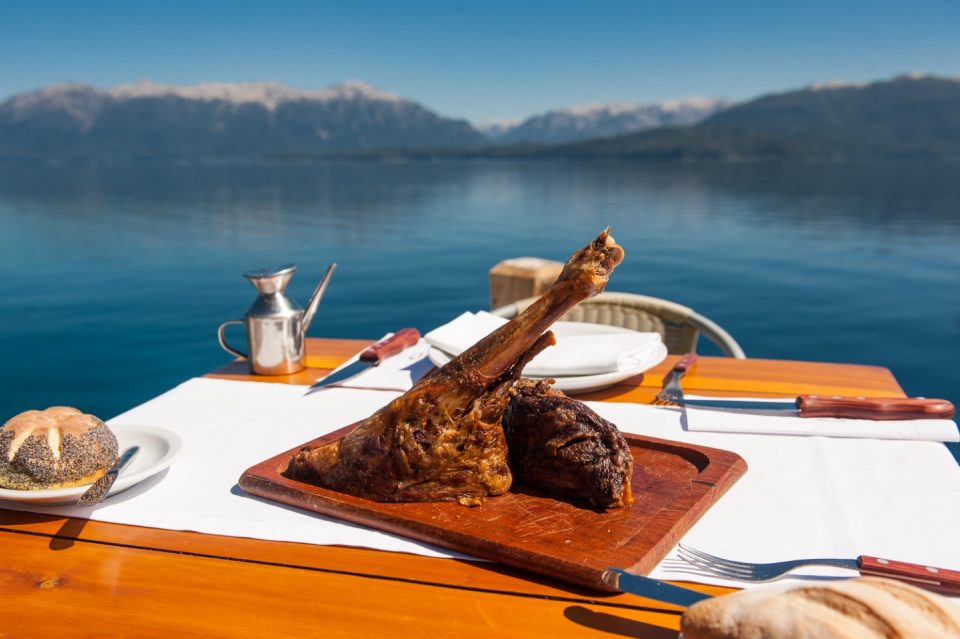 Una mesa con un plato con comida de asado y pan con el lago y montañas de fondo