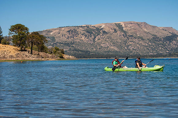 Dos hombreas andando en kayak con una costa con araucarias y montañas de fondo
